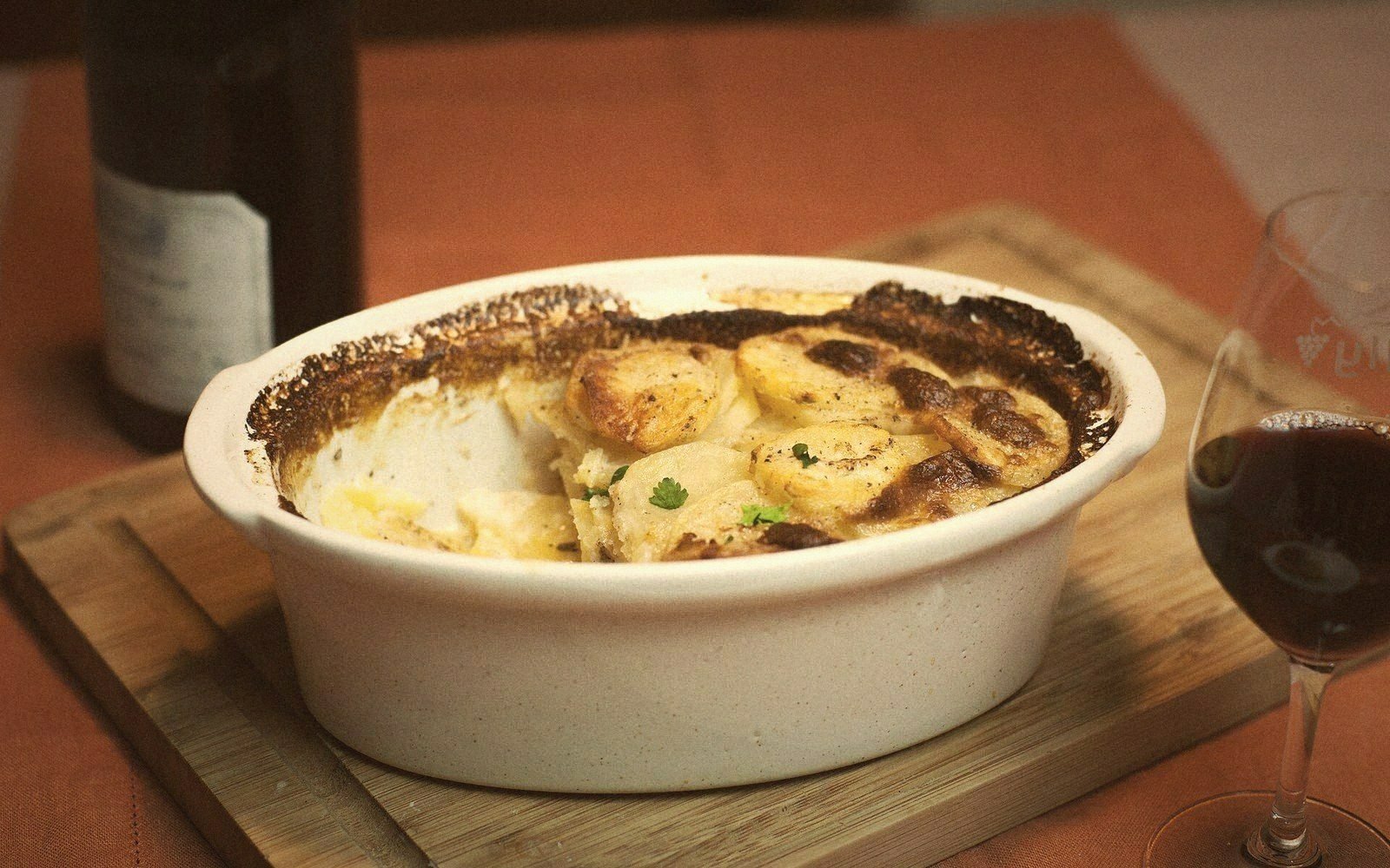 Baked potato gratin in a white oval dish beside a glass of red wine