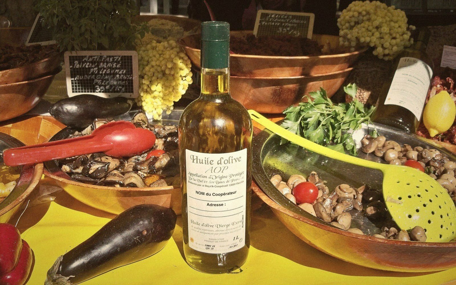 A bottle of AOP olive oil displayed among market bowls of vegetables, mushrooms, and grapes