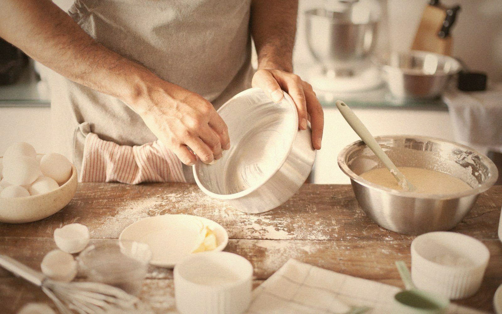 Hands greasing a baking dish beside bowls of batter, eggs and flour on a wooden counter