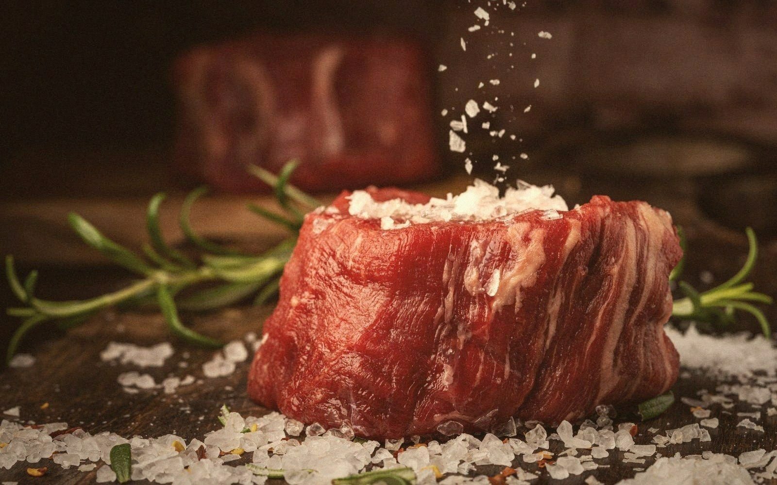 Coarse salt sprinkling onto a raw steak resting on rosemary sprigs and a wooden board