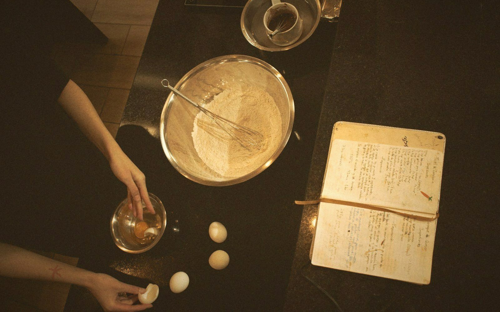 Overhead view of hands cracking eggs beside a flour bowl and handwritten recipe book